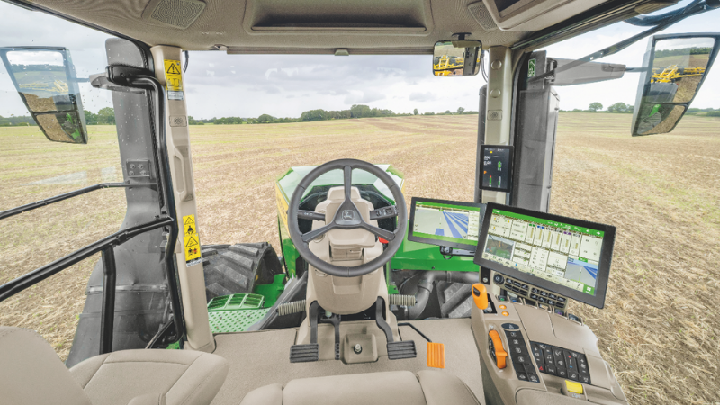 Vue intérieure d'une cabine de tracteur John Deere avec siège ergonomique beige, volant, écrans de contrôle et large pare-brise panoramique