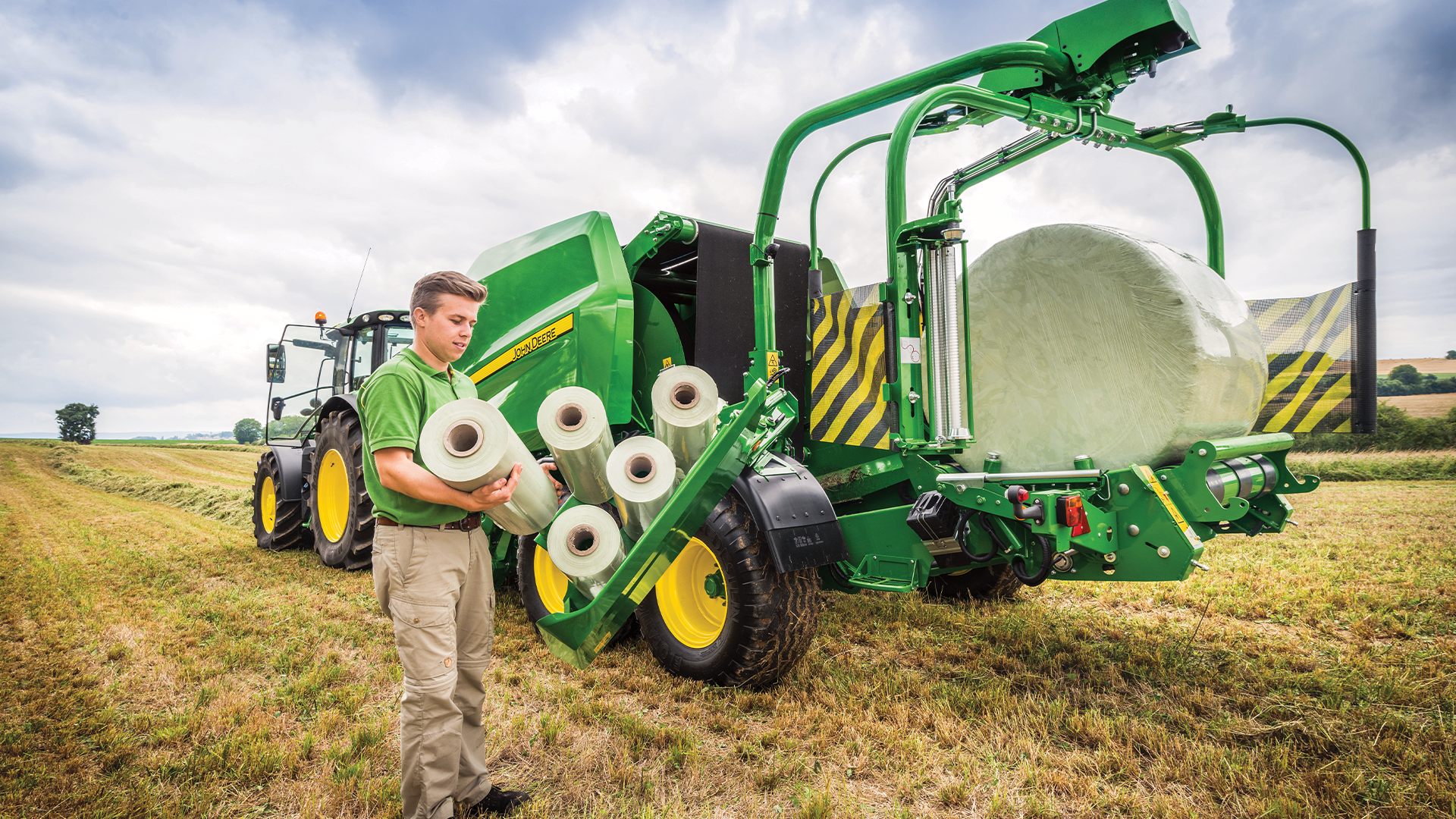 Agriculteur tenant des rouleaux de filets John Deere devant une presse à balles rondes en plein champ avec balles enrubannées