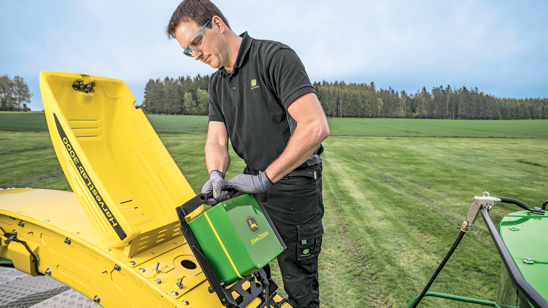 Technicien John Deere en polo noir inspectant une pièce détachée verte et jaune John Deere devant un équipement agricole jaune en plein champ, avec forêt de conifères en arrière-plan
