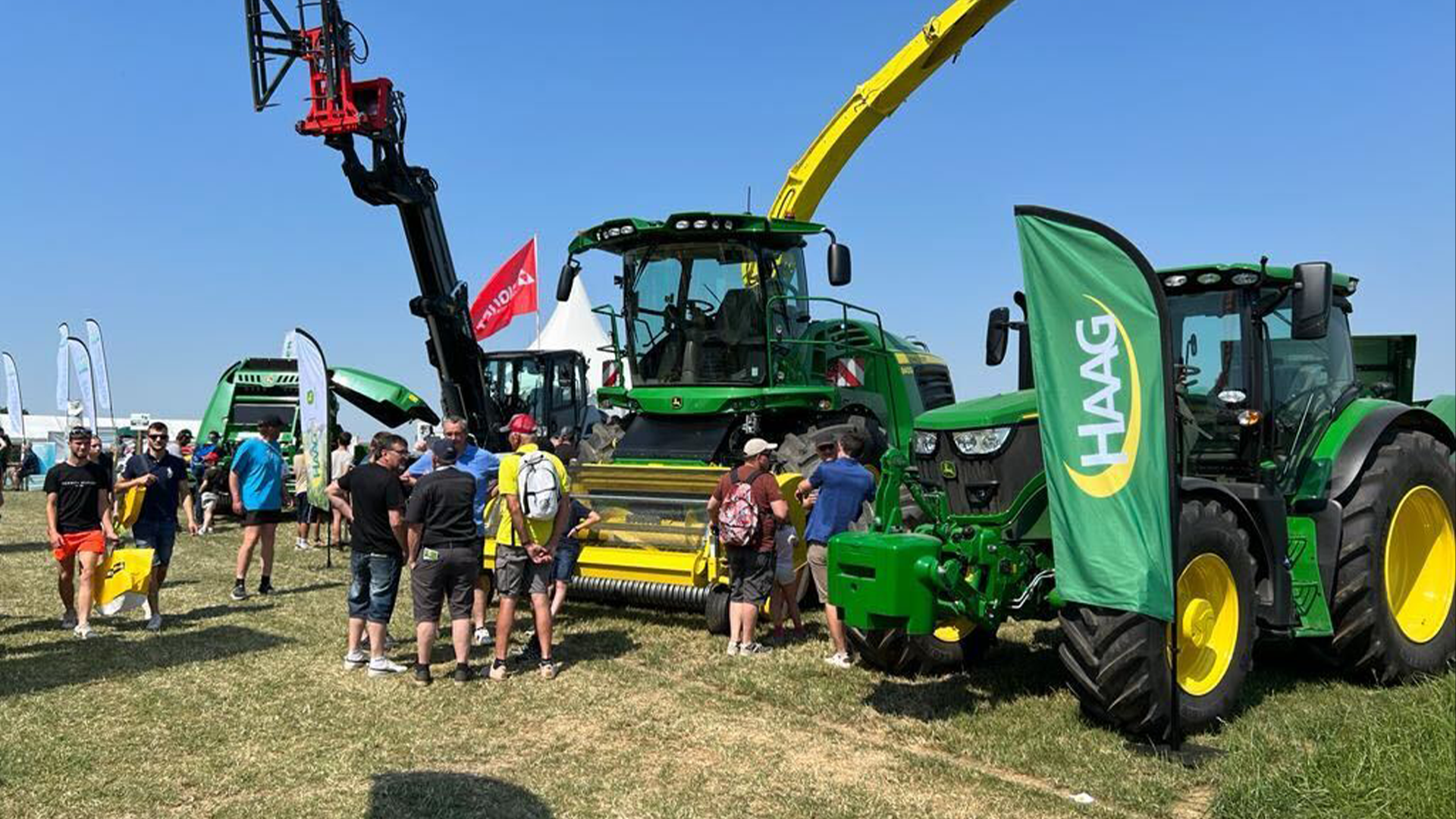 Visiteurs au Salon de l'Herbe 2026 autour de machines agricoles vertes et jaunes en démonstration en plein air, avec drapeaux HAAG, par temps ensoleillé