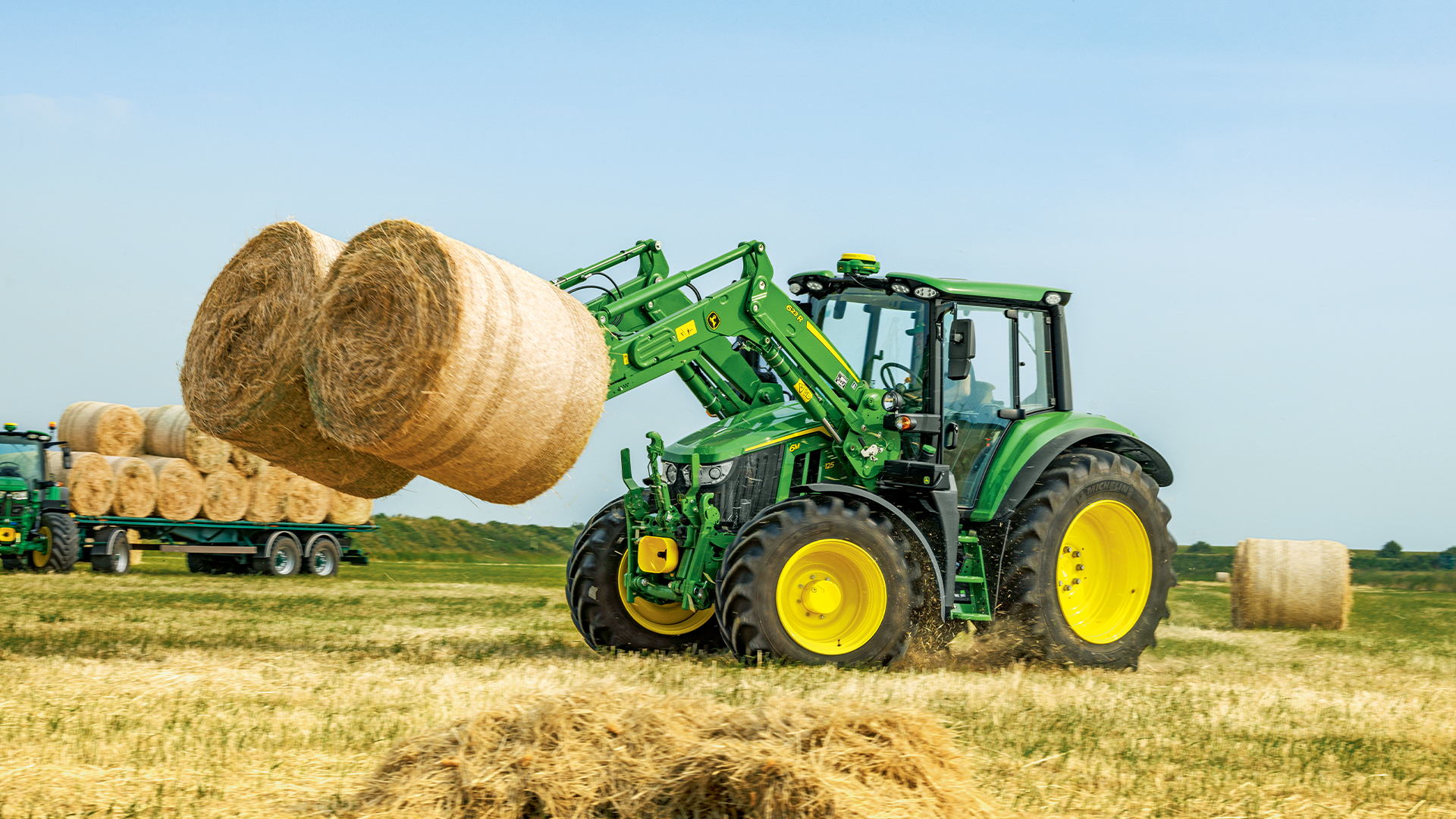 Tracteur John Deere avec chargeur frontal soulevant des balles de paille dans un champ après moisson.