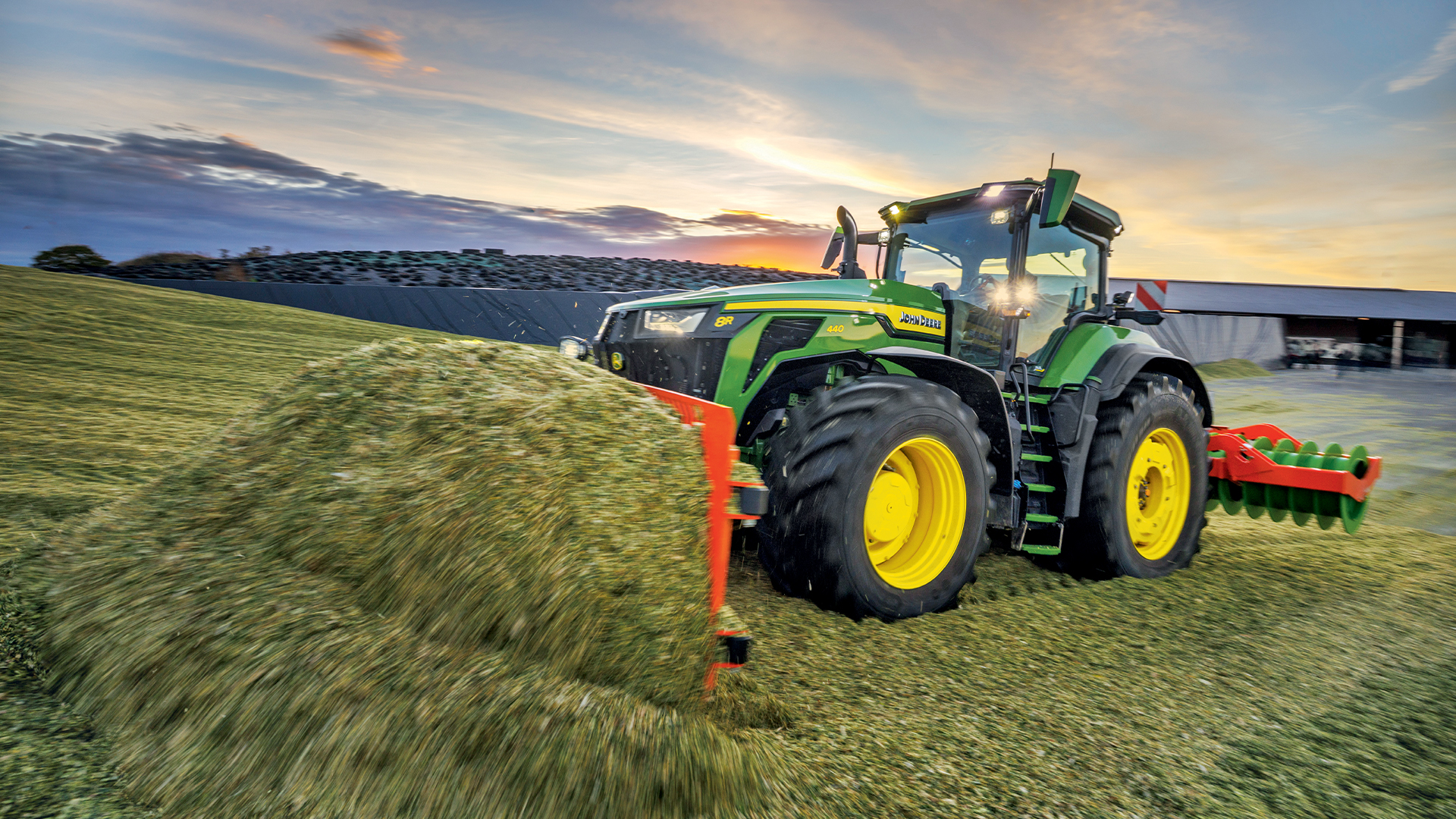 Tracteur John Deere 8R en action au crépuscule, travaillant une parcelle d'ensilage.